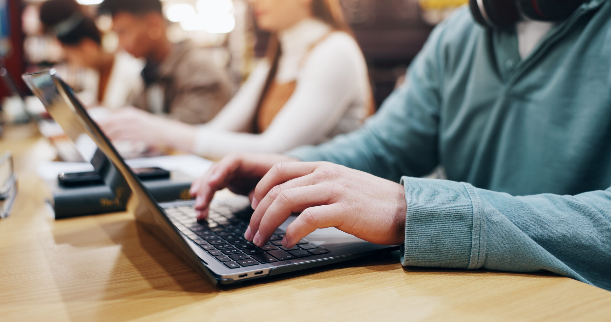 Close-up of a student typing on a laptop, with other students working at a shared table in the background.